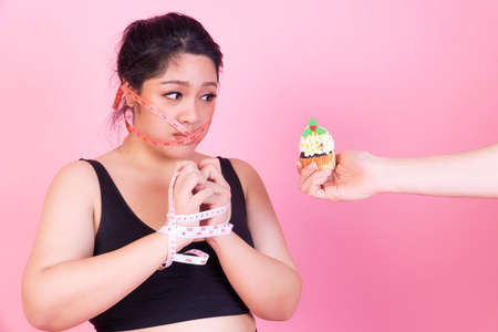 Diet restricts, weight loss, eating disorder concept. Obese overweight fat woman with measure tape cover her mouth while hands of man holding cupcake her thinking eat it over isolated pink background.の写真素材