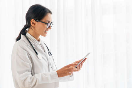 Friendly smile woman doctor or nurse wearing uniform and eyeglasses with stethoscope looking touchscreen tablet on own hand while standing beside windows at hospital. Healthcare technology concept.の写真素材