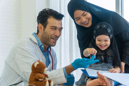 Health care concept. Muslim mother wear hijab with own little daughter visit professional doctor man while child sitting on the table with teddy in clinic. Man with stethoscope playing patient child.の写真素材