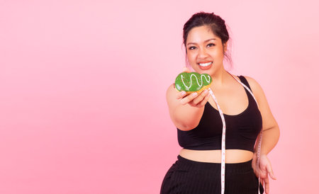 Obese unhealthy sweet junk food concept. Overweight young woman wear sport wear with measure tape holding or eating doughnut in own hand while standing over isolated pink background.の写真素材