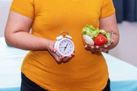 Healthy eating weight loss concept. Hand of fatty overweight woman holding alarm clock and glass bowl salad vegetable with fresh tomatoes and lettuce while standing at home.の写真素材