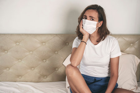 Quarantine at home concept. Unhappy young woman wear casual cloth and protective face mask worried something while sitting alone on white bed in the room.の写真素材