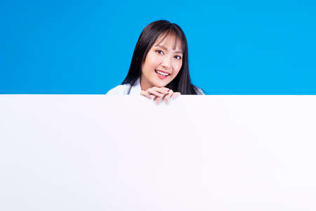 Banner, advertisement, sign board presentation concept. Positive cheerful young woman showing white placard empty copy space while standing over isolated blue background.の写真素材