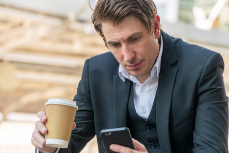 Lifestyle business success technology concept. Handsome manager businessman holding cup of coffee looking message in smartphone own hands while sitting over blur skyscraper city background.の写真素材