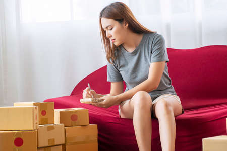 Startup sme e-commerce and new normal concept. Owner business young woman holding pen in own hand looking and writing customer order on notebook while sitting on red sofa with packaging box at home.の写真素材