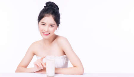 Charming lovely slim young woman with glass fresh milk beverage for good healthy skin while sitting on white table over isolated white background. Health care calcium food for fit body concept.の写真素材