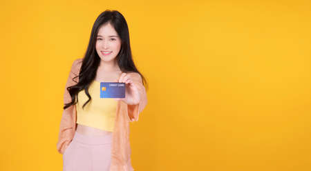 Cheerful asian woman showing credit card mockup standing with copy space over isolated yellow...