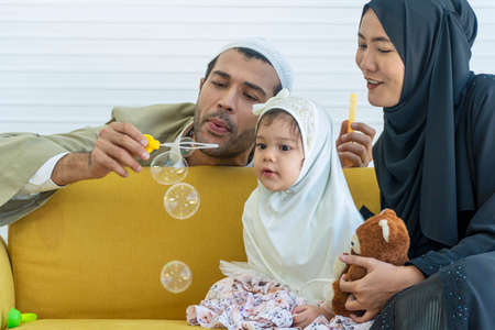 Enjoy mix race daughter wear head scarf with mother and father blowing bubbles shampoo while sitting...