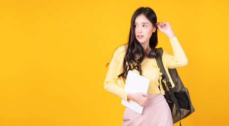 Beautiful college Asian woman holding tablet with backpack on yellow background. Student university young girl wear casual cloth with bag gadget tablet standing over isolated. Education campus conceptの写真素材