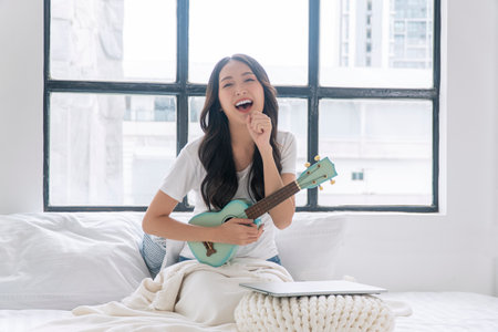 Cheerful asian woman playing ukulele sitting on white bed with pillows beside window at apartment. Joyful young girl holding acoustic ukulele in morning at home. Relax female play music ukulele hobby.の写真素材