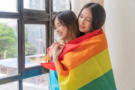 Joy romantic LGBT asian lesbian woman embracing girlfriend from back standing together beside window with rainbow flag at home. Smile asian young couple cuddle together with flag. LGBT relationshipの写真素材