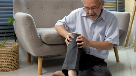 Elderly asian man touching knee hurt painfully problem sitting on floor at home. Patient older asian male injury leg disease mature knee suffering muscle sitting alone beside sofa. Physical therapy.の写真素材