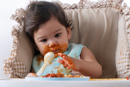 Adorable messy little child girl hungry use hand eating spaghetti sitting in high-powered chair at home. Toddler child with tomato sauce making mess face looking at food. Self-feeding conceptの写真素材