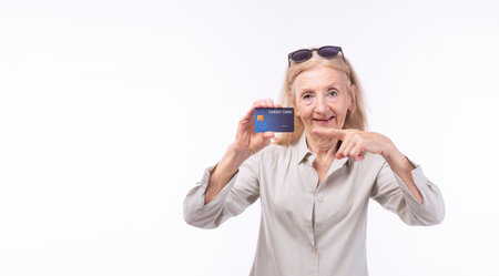 Happy friendly elderly woman with sunglasses showing credit card for payment standing over isolated...
