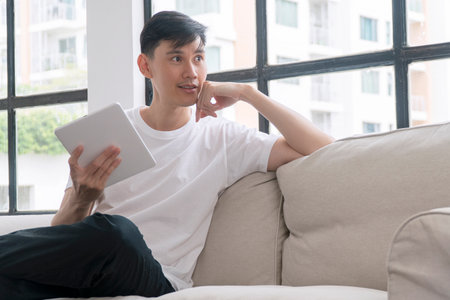 Handsome asian casual man holding digital tablet looking window thinking hand under chin sitting on...