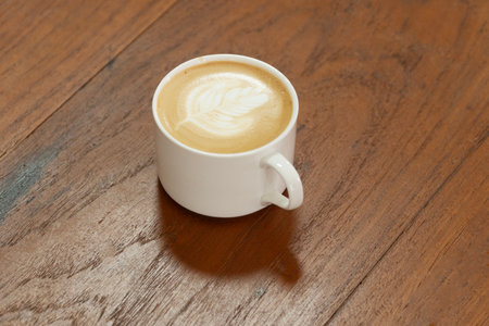 Mug cup of hot cappuccino art froth cream making flower on wooden table. Cup hot coffee fresh drawing pattern flower shaped milk froth foam on wood table. Beverage morning hot coffee art milk top vの写真素材