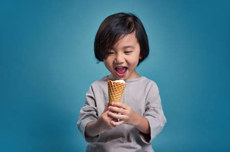 Excited asian little kid holding delicious ice cream. Empty space in studio shot isolated on colorful blue background. Education concept for school.の写真素材