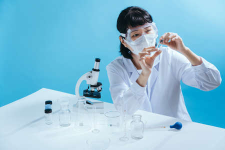 Smart young asian female scientist in lab coat with Medical face mask. Inspection on laboratory tools. Empty space in studio shot isolated on colorful blue background. Medical concept.の写真素材