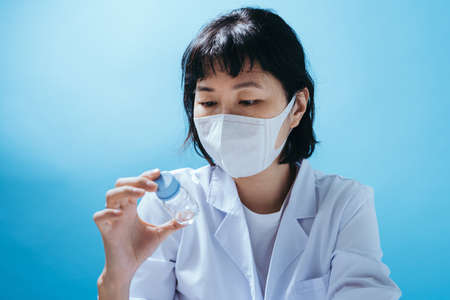 Close up smart young asian female scientist in lab coat with Medical face mask. Inspection on laboratory tools. Empty space in studio shot isolated on colorful blue background. Medical concept.の写真素材