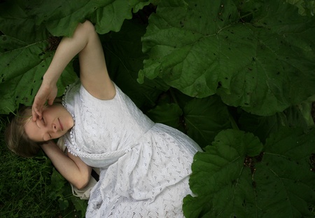 Young woman with white dress lying in garden between huge green leaves and enjoying nature.の写真素材