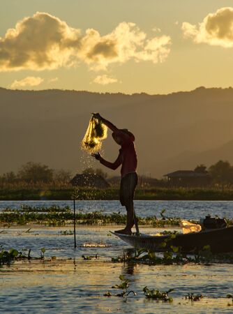 MANDALAY, MYANMAR. November 27, 2016. Against the background of the setting sun, the fisherman of Inle lake arranges the nets, standing on the edge of the boat, while simultaneously controlling the boat, holding the oar with his foot.のeditorial素材