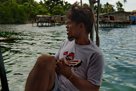 Semporna. Malaysia. November 30, 2018. A young resident of the island of Borneo plays the guitar and sings songs on the pier of the fishing village of sea gypsies, located on the shore of a reef islanのeditorial素材