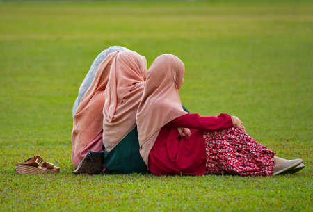Malaysia. Kuala Lumpur. Four young Muslim girls in beautiful pink hijabs relax on a grass lawn in...