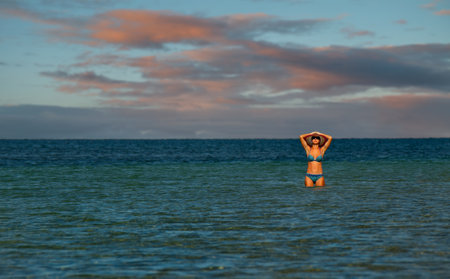 Crimea. Russia. August 10, 2018. A girl in a beach swimsuit and a hat standing in the water is sunbathing in the rays of the setting sun against the background of the endless horizon of the Black Sea.