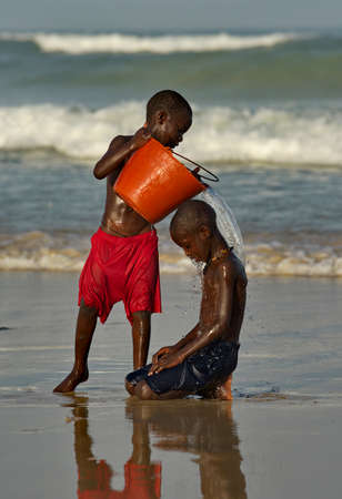 Saint-Louis. Senegal. October 11, 2021. Two village boys are doused with seawater from a plastic bucket on the sandy shore of the Atlantic Ocean.のeditorial素材