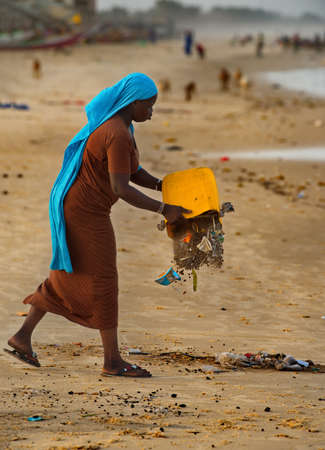 Saint-Louis. Senegal. October 10, 2021. A black village girl pours household garbage out of a plastic bucket on the shore of the Atlantic Ocean. That's how the oceans get clogged.のeditorial素材