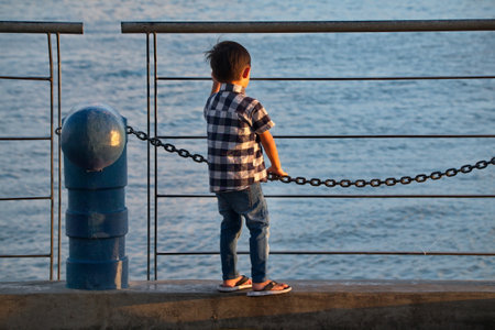 Semporna. Malaysia. A little boy stands near the fence of the city embankment and looks at the waves of the ocean, illuminated by the rays of the setting sun.の写真素材