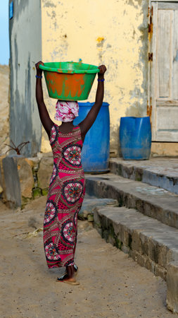 West Africa. Senegal. October 11, 2021. A young girl in national clothes gracefully carries a plastic basin with a load on her head along the street of the city of Saint-Louis.のeditorial素材