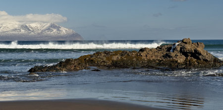 Russia. Far East, Kuril Islands. View of the cold waves of the Pacific Ocean against the background of snow-capped volcanoes surrounded by basalt rocks.の写真素材