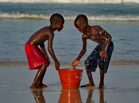 Saint-Louis. Senegal. October 11, 2021. Two village boys are doused with seawater from a plastic bucket on the sandy shore of the Atlantic Ocean.のeditorial素材