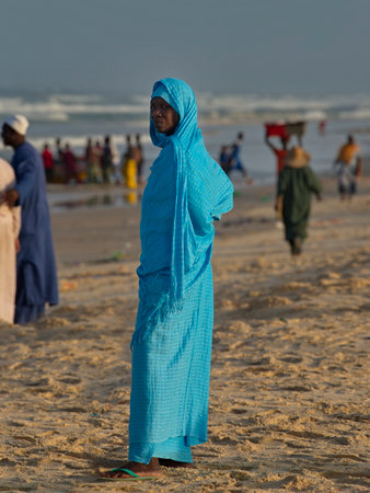Saint Louis, Senegal. October 11, 2021. Portrait of an African woman in a turquoise national dress against the background of the bustle of people on the Atlantic Ocean.のeditorial素材