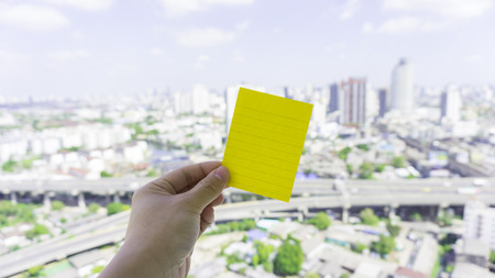 Green post it note put on urban scene with sky by a hand