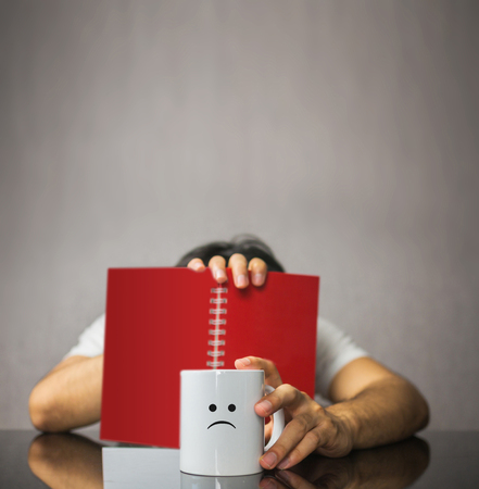 Sad face icon on coffee mug with man covering face with notebookの写真素材