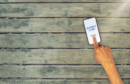 Enter Elderly Care app with senior hand touching the mobile screen on wooden tableの写真素材