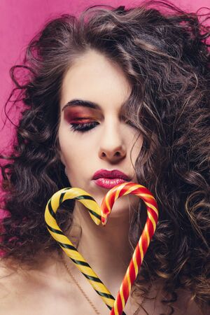 Curly beautiful girl with yellow and red lollipop sweet stick on pink backgroundの写真素材