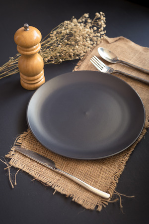 Black cutlery set - knife, spoon, fork, on black plate on sackcloth and dark wooden table. Top view.の写真素材