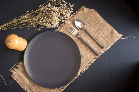 Black cutlery set - knife, spoon, fork, on black plate on sackcloth and dark wooden table. Top view.の写真素材