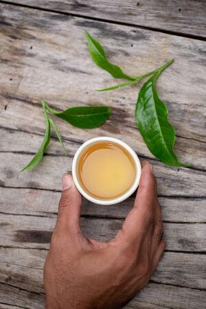Glass of tea in hand on a wooden table and the top of fresh tea leaves.の写真素材