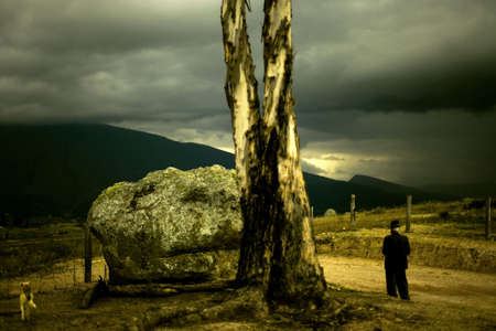 A traditional colombian farmer and his dog walk past a large rock and tree in the imposing colombian countryside with huge mountains and a turbulent sky in the background.の写真素材