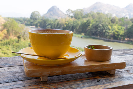 Yellow coffee cup and coffee spoon on wooden table with scenic mountain and river view.の写真素材