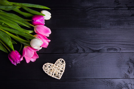 Beautiful bouquet of tender pink and white tulips with white wicker heart on black wooden background.の写真素材