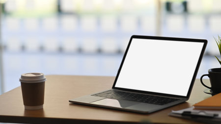 Photo of Computer laptop with white blank screen putting on wooden working desk with takeaway coffee cup, clipboard and notebook over modern cafe as background. Orderly/Comfortable workplace concept.の写真素材