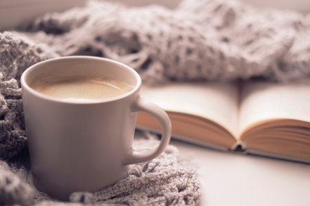 Cozy home with cup of coffee and a book. Hygge style. Mug of black coffee wrapped in warm scarf on wooden board.の写真素材