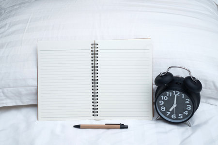 Diary or notebook and vintage alarm clock on bed in bedroom at home or hotel, working or note something before sleep.の写真素材
