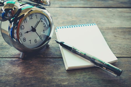 Time management concept: vintage tone alarm clock, pen and notepad on old wooden table
