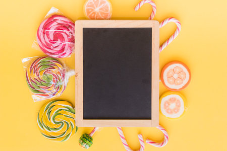 top view of blackboard with candies and lollipops on yellow background